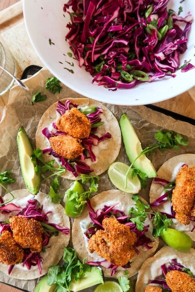 Overhead shot of crispy cod tacos with pickled slaw and chilli crema on a white plate with lime wedges.